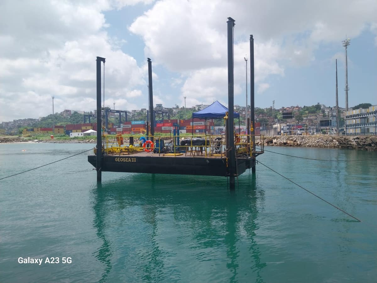 Balsa auto-elevatória GEOSEA III atracada nas águas de um porto, com os quatro spuds erguidos e um toldo azul montado sobre o convés.
